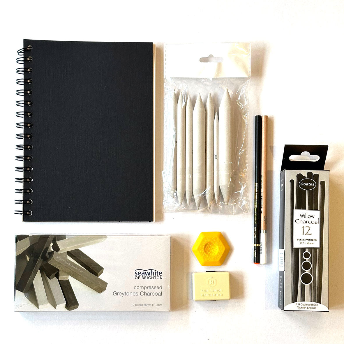 The Charcoal Kit Box containing a collection of art supplies including a black notebook, charcoal sticks, and packaging on a white background