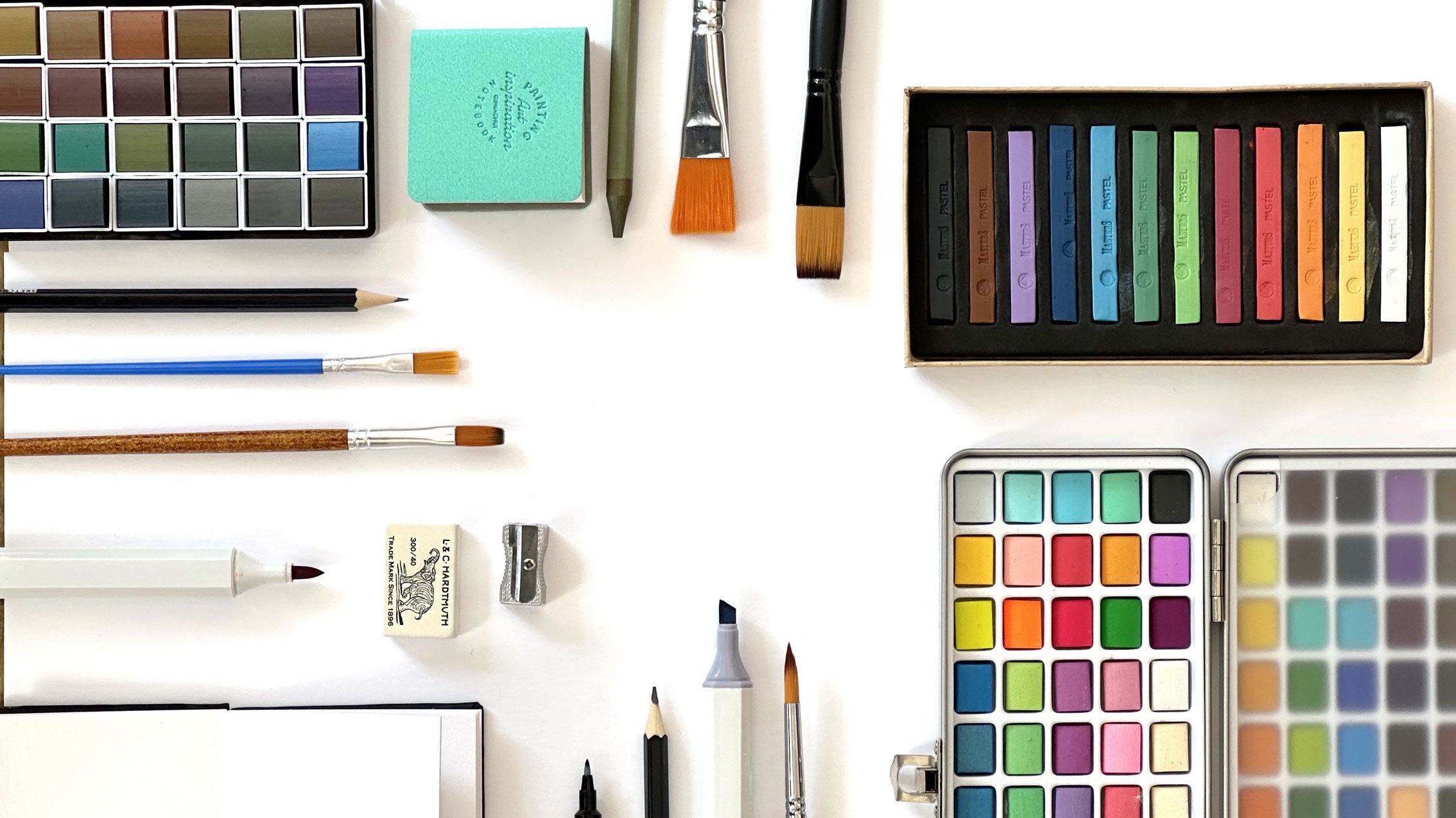 Assorted art supplies including paintbrushes, watercolour paints, pastels, sketchbooks and pens.
